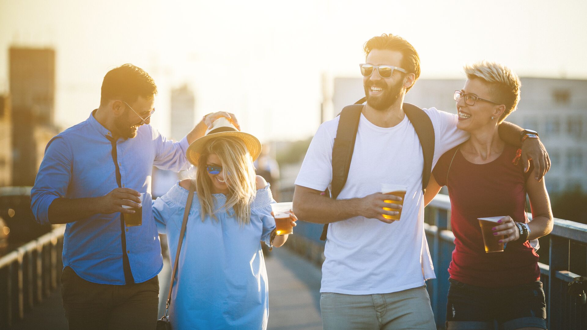 Walking Beer Hipster people are walking through the bridge. This group of friends are holding plastic glasses with beer in it and drinking it. They are enjoying at amazing sunset behind them. There are two men and two women who are spending great time together. It's weekend and they decide to relax and hanging out together on traveling. Everybody are dressed casual and comfortable so they are walking a lot this afternoon.