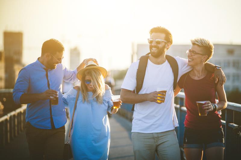 Walking Beer Hipster people are walking through the bridge. This group of friends are holding plastic glasses with beer in it and drinking it. They are enjoying at amazing sunset behind them. There are two men and two women who are spending great time together. It's weekend and they decide to relax and hanging out together on traveling. Everybody are dressed casual and comfortable so they are walking a lot this afternoon.