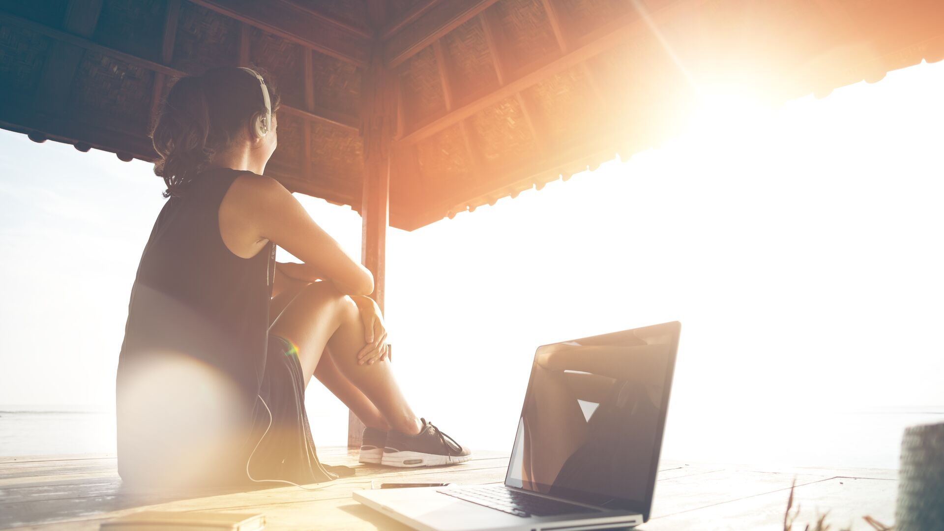 Laptop Outside Woman freelancer listening music at sunrise near ocean. Blurry effect, lens flares effect, intentional sun glare